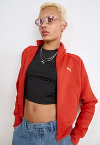 Veste zippée rouge avec des rayures blanches, portée sur un top noir court. Un jean bleu, des colliers en argent superposés et des lunettes de soleil roses complètent le look.