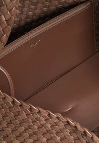 Brown woven bag featuring a smooth, brown leather interior with a zippered compartment and subtle logo detail on the leather.