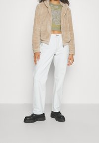 Beige, flauschige Jacke mit Reißverschluss und Kapuze, getragen über einem grünen, gemusterten Crop-Top. Hellblaue, gerade geschnittene Jeans und schwarze Plateauschuhe vervollständigen das Outfit.