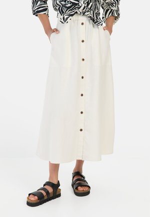 White button-up skirt with an elastic waistband, featuring brown buttons down the front and side pockets. Paired with black strappy sandals.