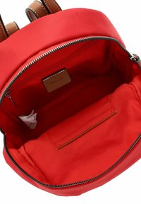 Intérieur d'un sac à dos en tissu rouge avec une texture lisse, présentant une poche zippée et une petite étiquette en cuir à l'intérieur.