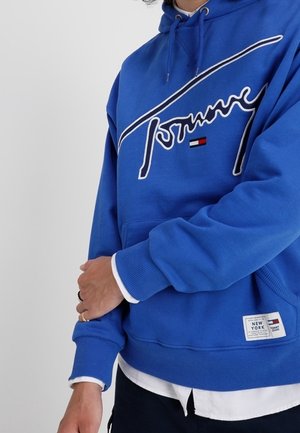 Personne portant un sweat à capuche bleu Tommy Jeans avec des cordons blancs, un t-shirt blanc en dessous et un pantalon noir.