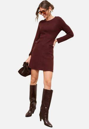 Rochie tricotată până la genunchi, cu model în relief, de culoare burgundă închisă, cu mâneci lungi și buzunare frontale. Asortată cu cizme strălucitoare, până la genunchi, cu model de croco.