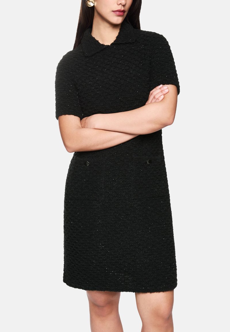 Schwarzes kurzärmeliges Kleid mit strukturierter Oberfläche und Kragen, versehen mit zwei dekorativen schwarzen Knöpfen vorne.