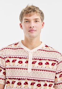Korte mouwen, crèmekleurig shirt met een ge puffte teddybeerpatroon in rood en beige, met een knopenlijst en een geometrisch achtergronddesign.
