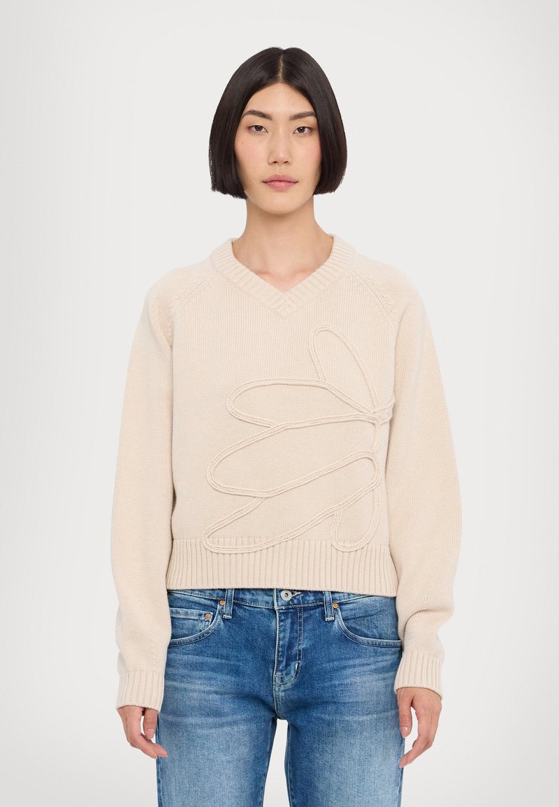 Pull en tricot beige avec un col en V, présentant un motif floral texturé prononcé. Manches et ourlet côtelés, associé à un jean bleu.