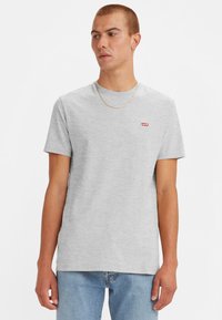 Tricou din bumbac gri, cu guler rotund, mâneci scurte și un mic logo roșu Levi's pe pieptul stâng. Purtat cu blugi albaștri deschiși.