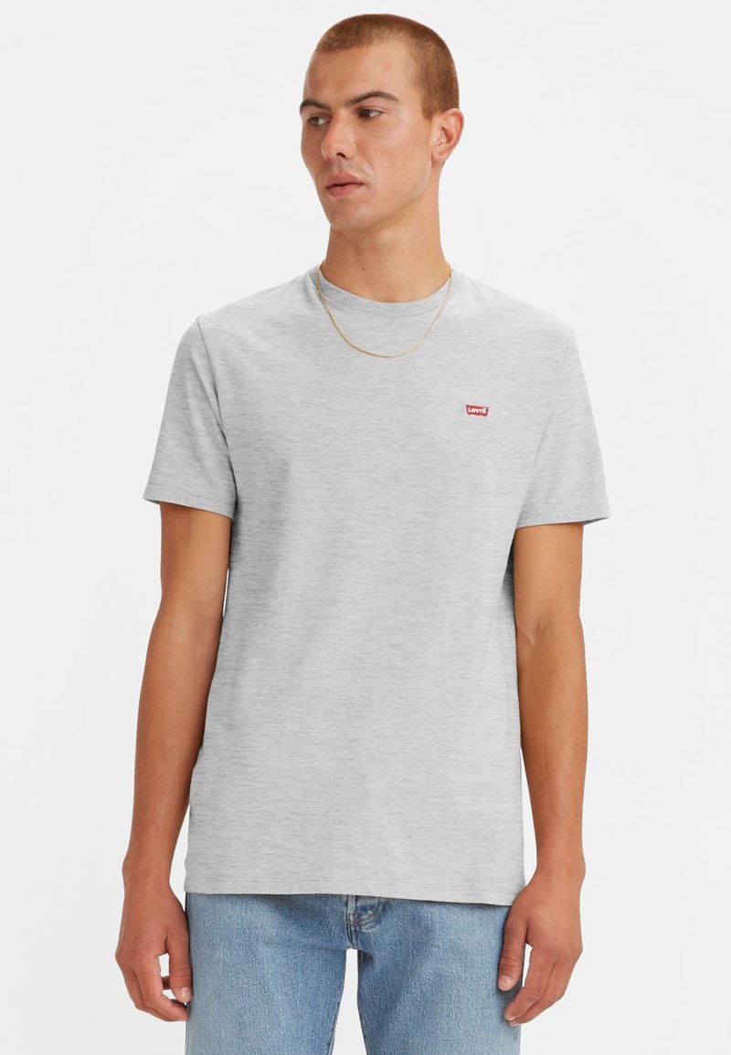 Tricou din bumbac gri, cu guler rotund, mâneci scurte și un mic logo roșu Levi's pe pieptul stâng. Purtat cu blugi albaștri deschiși.