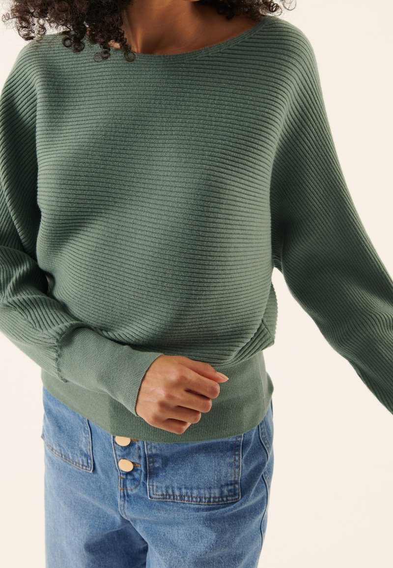 Femme portant un pull côtelé vert sauge avec col bateau et manches retroussées, associé à un jean bleu taille haute avec deux boutons sur le devant.