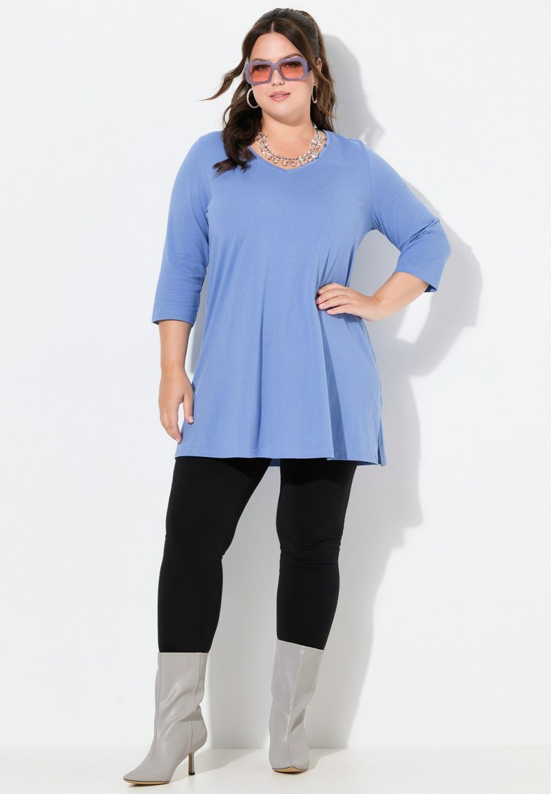 Blaue Tunika mit Dreiviertelärmeln, seitlichen Schlitzen, kombiniert mit schwarzen Leggings und grauen Absatzstiefeln; accessoriert mit einer Statement-Halskette.