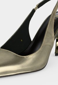 Gros plan d’une chaussure à talon haut slingback métallisée dorée avec une semelle intérieure noire et un talon bloc doré brillant.