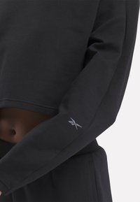 Černá zkrácená mikina vyrobená z měkké tkaniny, s kulatým výstřihem a malým šedým logem na levém rukávu.