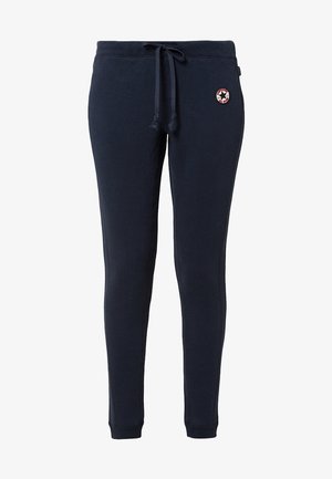 Mujer que lleva chaqueta de cuero negra sobre camisa azul y top blanco, pantalones azul marino con cordón y zapatillas deportivas negras y blancas con estampado.