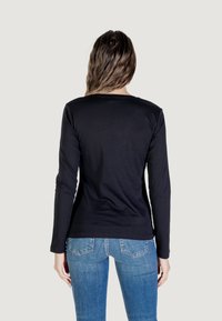 Donna con una maglietta nera a maniche lunghe e jeans blu, rivolta di spalle verso la fotocamera su uno sfondo grigio chiaro.