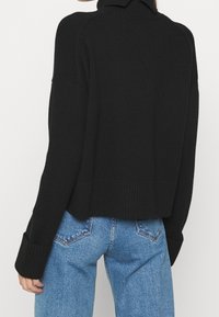Persona che indossa un maglione a coste nero con maniche lunghe e jeans in denim blu, mostrata di spalle contro uno sfondo bianco semplice.