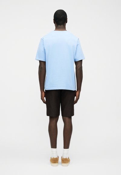 Άντρας που στέκεται γυρισμένος με την πλάτη, φορώντας ανοιχτό μπλε κοντομάνικο πουκάμισο, μαύρα σορτς, λευκές κάλτσες και ταμπα αθλητικά παπούτσια σε απλό φόντο.