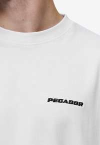 Camiseta de algodón blanca con cuello redondo y un pequeño logotipo bordado en negro "PEGADOR" en el lado izquierdo del pecho.
