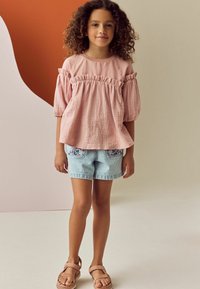 Hellrosa, strukturiertes Oberteil mit Rüschem Ärmeln, kombiniert mit pastellgestreiften Shorts mit bunten Taschenstickereien. Sandalen runden den Look ab.