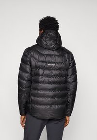 Giubbotto imbottito nero con un design trapuntato, dotato di cappuccio e logo "MAMMUT" sul retro. Texture liscia e leggera, vestibilità fino alla vita.