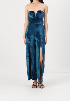 Femme portant une combinaison en velours bleu sarcelle sans bretelles avec des fentes à l’avant sur des jambes larges et des sandales fines noires sur un fond uni.