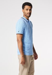 Světle modré polo tričko s krátkým rukávem, s texturovaným tkaným vzorem a kontrastním tmavým límcem. Rýhovaný lem a detail značky na rukávu.