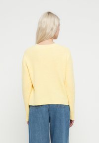 Pikajuukseline blond naine, vaadates selja tagant, seljas helekollane kampsun ja sinised kõrge vöökohaga teksad, taustal on lihtne valge sein.