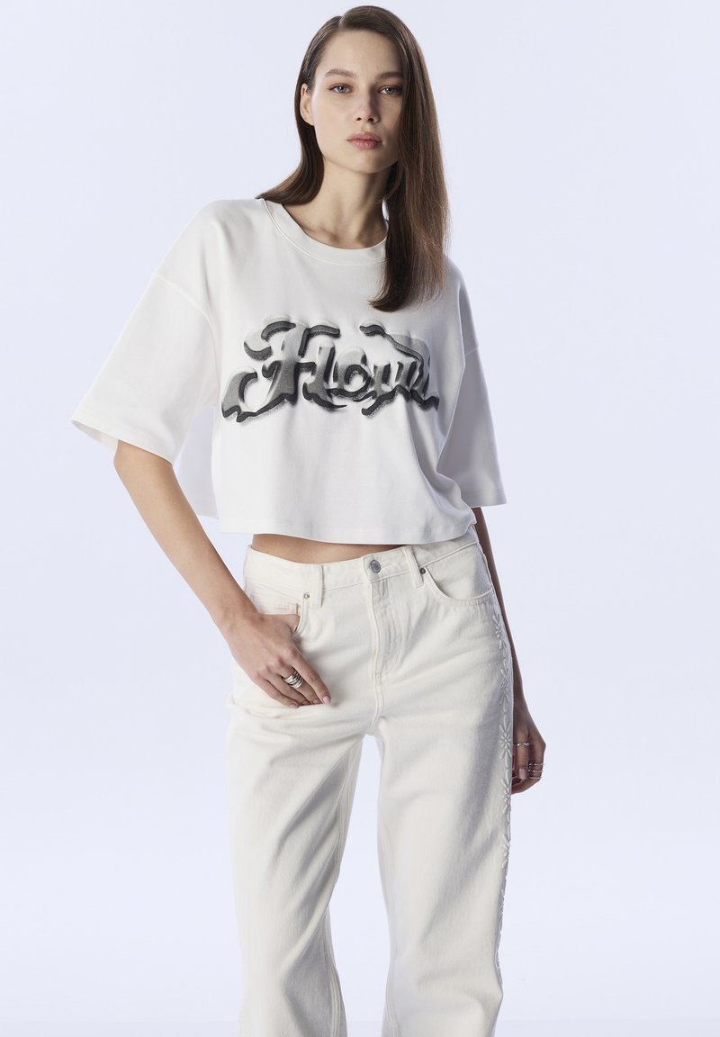 Zusammengetragener, weißer T-Shirt mit schwarzem Schriftzug "Floyd." Kombiniert mit hochgeschnittenen, hellen Jeans mit seitlichen Stickdetails.