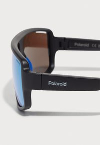 Occhiali da sole Polaroid neri con finitura opaca, lenti con sfumatura blu e dettagli del logo argentato sulle tempie. Design elegante e moderno.