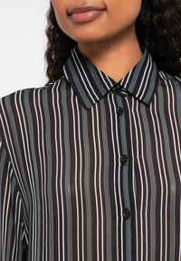 Close-up van een persoon die een zwart, grijs en wit verticaal gestreept overhemd met een puntkraag draagt.