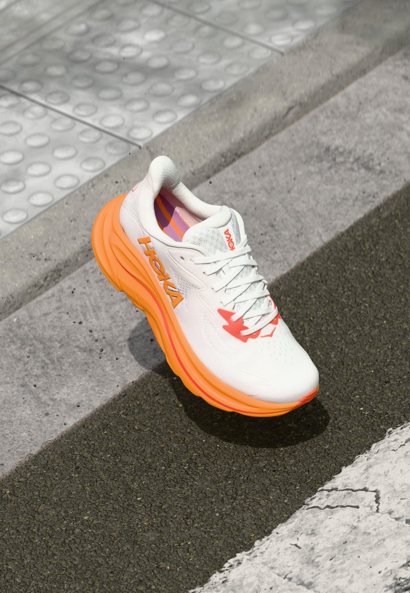Chaussure de course blanche avec des accents orange et rose. Tige en maille, semelle épaisse et rembourrée, et détail de logo bien visible. Semelle extérieure texturée pour une bonne adhérence.