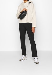 Sudadera beige con capucha, pantalones negros acanalados y zapatillas grises con detalles amarillos. Pequeño bolso bandolera negro con detalles de cremallera.