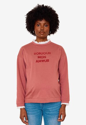 Woman with curly hair wearing a pink sweatshirt reading "Bonjour Mon Amour" and blue jeans, standing against a plain white background.