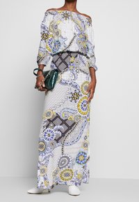 Robe longue à épaules dénudées présentant un motif floral et géométrique coloré, avec des manches longues et une coupe ample. Accessoirisée d'un sac à main vert.