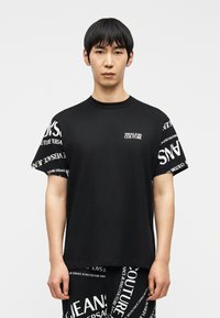 Νέος άνδρας που στέκεται προς τα εμπρός, φοράει μαύρο μπλουζάκι και παντελόνι Versace Jeans Couture με λευκά μοτίβα κειμένου της μάρκας.