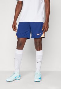 Nike modré športové šortky s oranžovými akcentmi, v kombinácii s bielymi vysokými ponožkami a svetlomodrými futbalovými kopačkami so š blackými detailami.