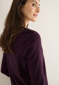 Langærmet, tætsiddende top i mørk lilla bomuld. Glat tekstur med minimale sømme. Visningen viser ryggen og skuldrene med løst, naturligt hår.