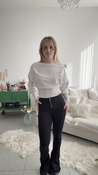 Νεαρή γυναίκα στέκεται σε φωτεινό σαλόνι φορώντας λευκό πουλόβερ και μαύρο παντελόνι, με ορατή πράσινη ντουλάπα, καναπέ και χνουδωτό χαλί.