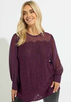 Blusa viola con un fronte in pizzo e maniche lunghe trasparenti. Motivo floreale testurizzato sul pizzo. Collo arrotondato con un delicato bordo.