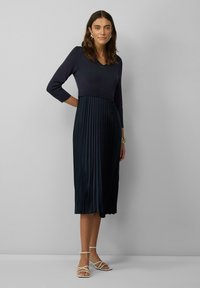 Marineblaues Plisseekleid mit einem enganliegenden, gerippten Oberteil, Dreiviertelärmeln und Midi-Länge. Getragen mit weißen Riemchensandalen.