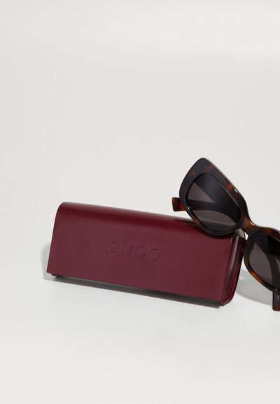 Cutie de ochelari Gucci din piele bordo, cu logo embossat, conținând ochelari de soare supradimensionați cu montură din chihlimbar și lentile întunecate, cu o textură netedă.