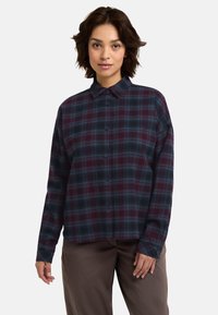 Camicia a quadri rossa e blu con colletto, maniche lunghe e vestibilità comoda. Presenta piccoli bottoni e una texture di tessuto morbido.