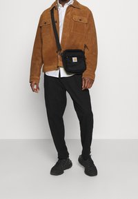 Muž v hnědé semišové bundě, bílém tričku, černých kalhotách, černých teniskách a malé černé kabelce přes rameno s logem na přední straně na bílém pozadí.