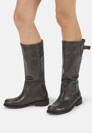 Dunkelbraune Lederstiefel bis zur Mitte der Wade mit seitlicher Schnalle, getragen an nackten Beinen vor einfarbigem Hintergrund.