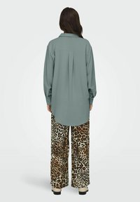 Long-sleeve shirt in muted teal, featuring a button-down collar and a back pleat, paired with wide-leg pants in a beige and brown leopard print.