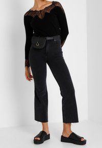 Haut noir à manches longues avec des détails en dentelle, jeans foncés taille haute et sandales noires à enfiler. Petite sacoche noire avec une boucle circulaire.