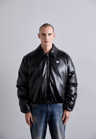 Mann mit kurzem Haar, der eine schwarze Lederjacke mit Reißverschluss und einem kleinen Metalllogo auf der Brust sowie blaue Jeans trägt, vor einer schlichten Wand.
