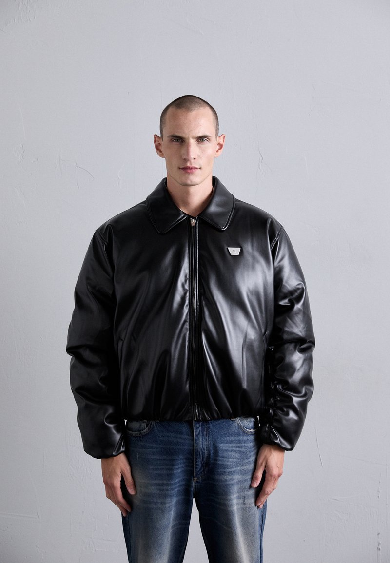 Homme avec les cheveux courts portant une veste en cuir noire zippée avec un petit logo en métal sur la poitrine et un jean bleu devant un mur uni.