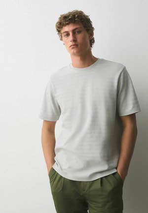 Młody mężczyzna z kręconymi włosami, ubrany w jasnoszarą teksturowaną koszulę z krótkim rękawem i oliwkowozielone spodnie, z rękami w kieszeniach, stojący przy białej ścianie.