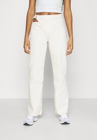 Pantalons larges couleur crème avec une finition texturée, présentant une taille diagonale unique et un accent en boucle métallique.