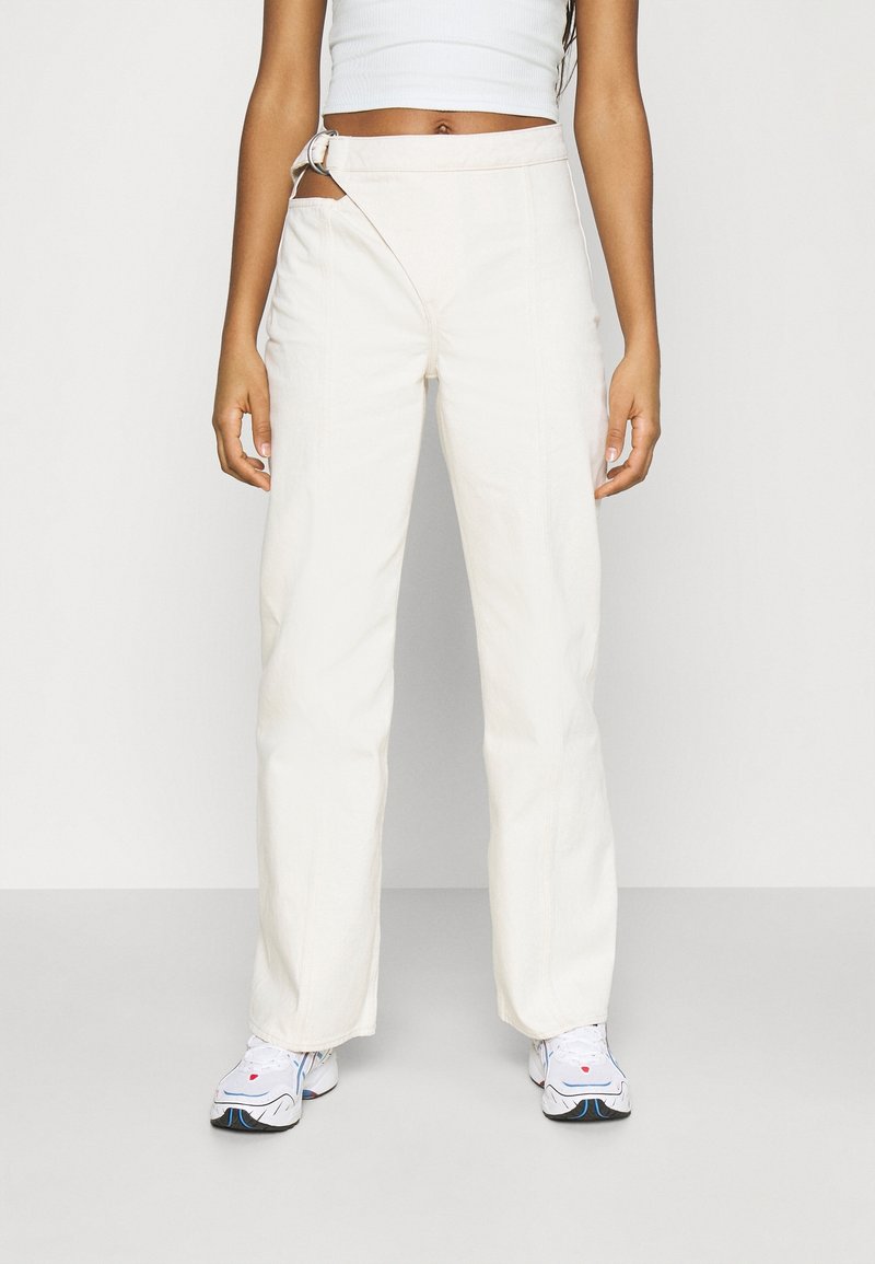 Pantalons larges couleur crème avec une finition texturée, présentant une taille diagonale unique et un accent en boucle métallique.